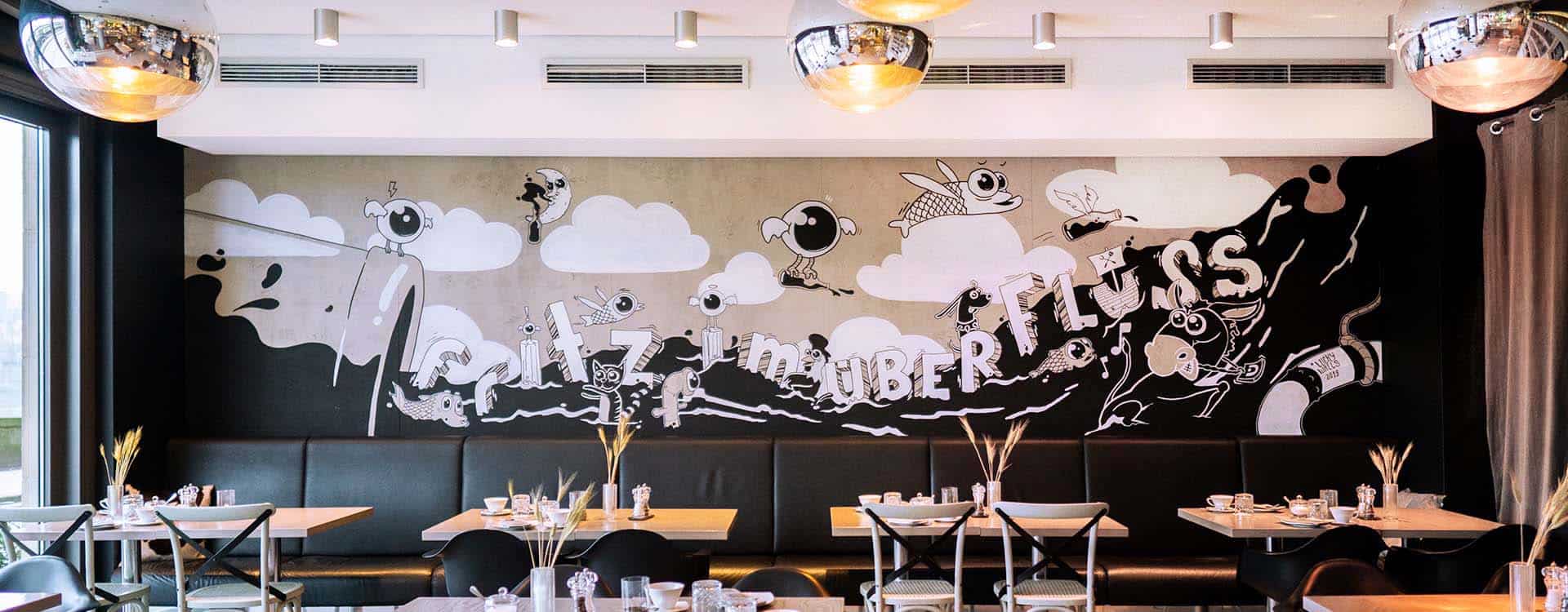 Großflächiges Wandbild in einem Restaurant mit surrealen, schwarz-weißen Illustrationen und verspielten Schriftzügen.